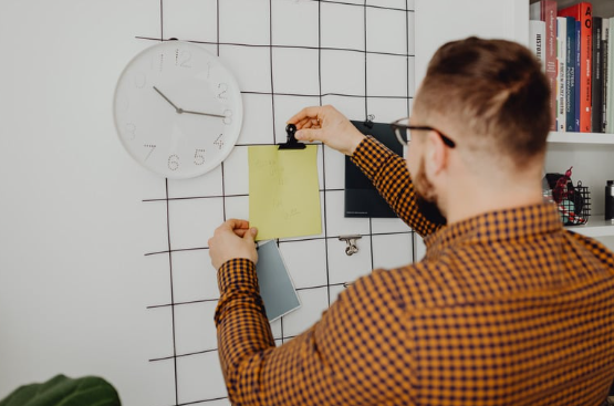 Man in a checkered shirt pins a yellow note on a gridded wall beside a white clock in a bright office.