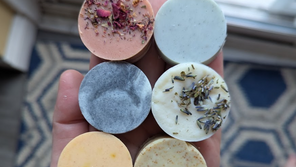 Hand holds six round soap bars in pastel colors, some topped with dried flowers and lavender, near a window over a blue patterned rug.