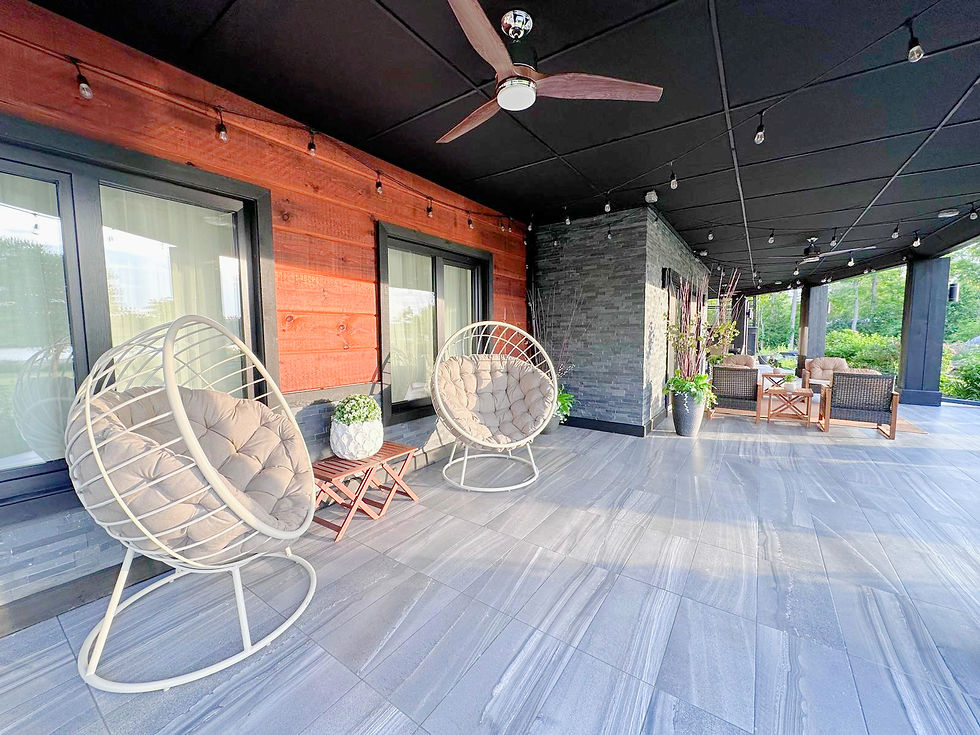Modern covered patio with hanging egg chairs, cushioned seating, string lights, and gray tile floor beside wood and stone walls.