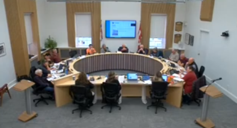 People seated around a circular table in a meeting room, screen on wall displaying presentation, Canadian flags visible, calm atmosphere.