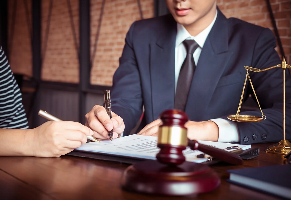 Lawyer in dark suit signs contract at desk with client, gavel and scales of justice in foreground, brick office background.
