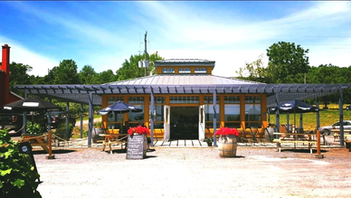 Sunny outdoor café with a wooden building, patio tables, black umbrellas, red flower barrels, and a chalkboard menu board.