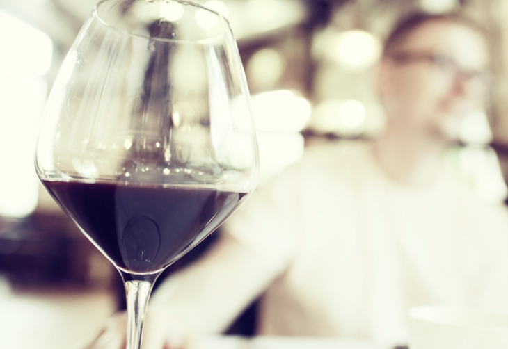 Close-up of a wine glass with red wine in a softly lit restaurant, with a blurred person in the background.