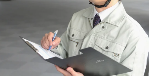 Person in a light gray work jacket holding a pen and clipboard, writing notes. Indoor setting with a neutral background. Calm atmosphere.