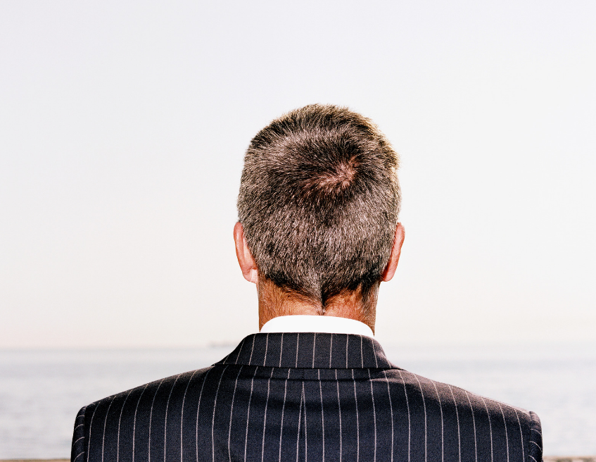 Back view of a man in a pinstripe suit looking out over the sea under a bright, blank sky.