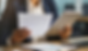 Businessperson in blazer reviews a printed document and tablet at an office desk, with a laptop and chart papers nearby.