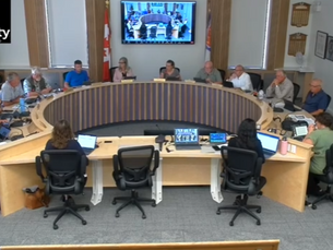 A group of people sit around a U-shaped conference table, engaging in discussion. Screens display meeting content. Walls have plaques and flags.