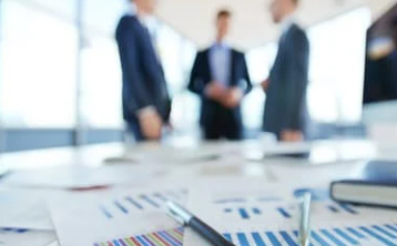 Blurred image of three people in suits discussing, standing in a bright office. Focus on close-up documents with graphs on a desk.