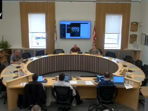 Panel of five people in a curved wooden-seated room, using laptops. Screen displays a video call. Flags outside windows. Warm lighting.