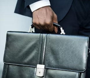 A suited person grips a black leather briefcase in a blurred indoor setting, suggesting a formal business mood.