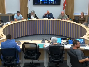 People sit around a curved conference table with laptops and papers, engaged in discussion. Flags and framed items decorate the wood-paneled room.