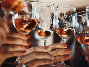 Hands with painted nails clinking wine glasses filled with rosé in a celebratory toast. Warm lighting enhances the festive mood.