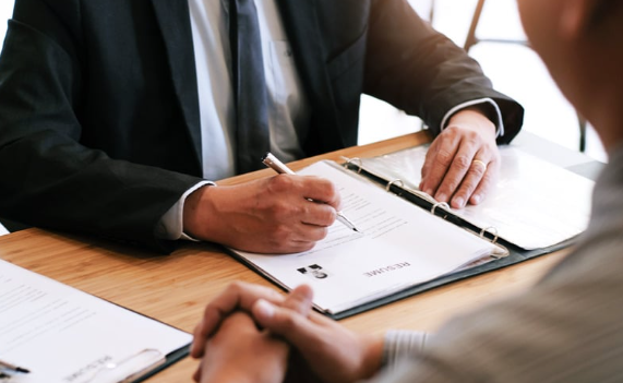 Business meeting with a man in a suit writing on paperwork at a table while another person listens with clasped hands.