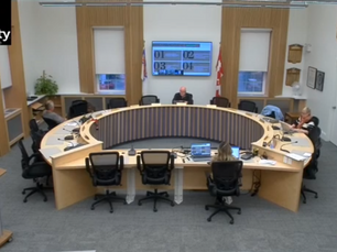 Meeting in a circular conference room with five people, screens displaying numbers, office furniture, and flags. Text reads "The County."