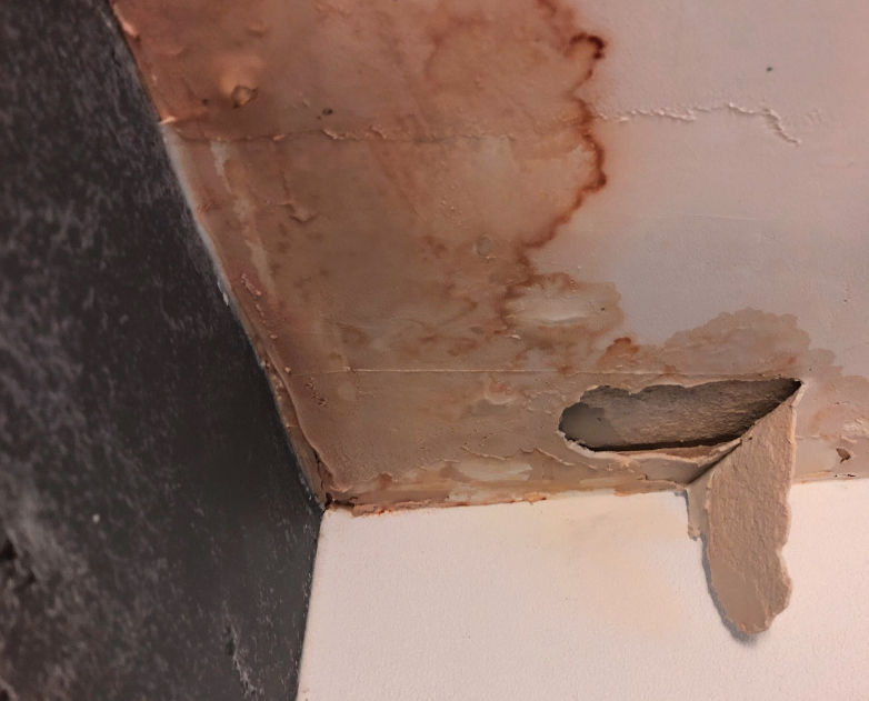 Water-damaged ceiling corner with peeling plaster, brown stains, and a hole exposing damaged material above a white wall.