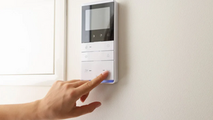Hand pressing a white wall-mounted touchscreen control panel in a bright room.