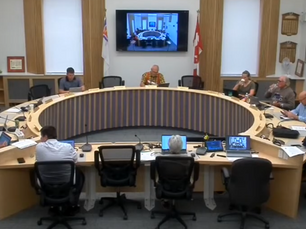 People seated around a large circular table in a conference room, with laptops open. A screen displays a similar room. Flags and plaques adorn the walls.