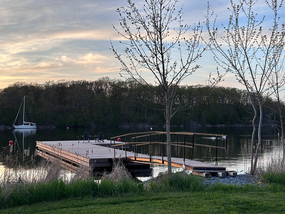Tranquil lakeside scene with a wooden dock leading to calm water, framed by budding trees. A sailboat is moored in the distance, evoking serenity.