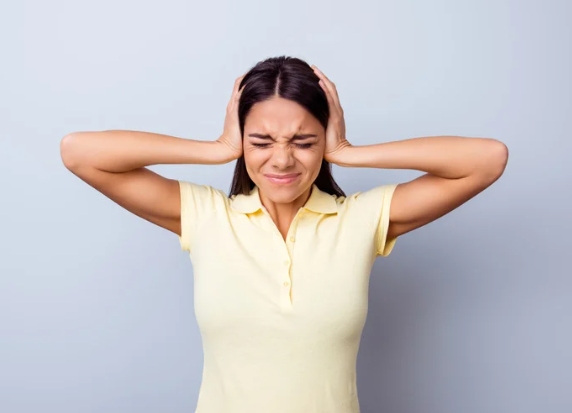 Woman in a yellow shirt holding her head with closed eyes, expressing frustration. Background is plain light gray.