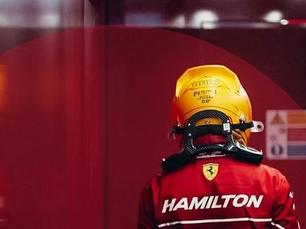 Lewis Hamilton in Ferrari race suit and yellow helmet, seen from behind, walking through the team’s garage area.