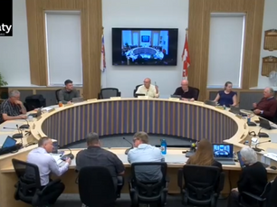 Meeting in progress around a circular table in a conference room. People are engaged in discussion. A screen displays the same scene.