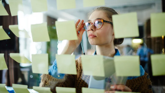 Woman in glasses adjusts her frames behind a glass wall covered with yellow sticky notes in a bright office, looking focused.