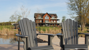 Two gray Adirondack chairs on a wooden dock by a pond, with a rustic house and trees in the background on a calm day
