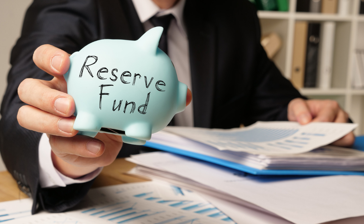 Businessman holds a light blue piggy bank labeled Reserve Fund over office charts and papers on a desk.