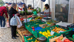 Customers browse a busy outdoor produce market, with an elderly vendor serving fruit in crates under a tarp.