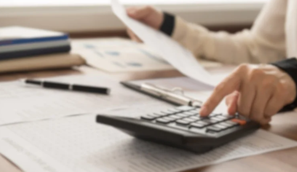 A person uses a calculator on a wooden desk with papers, a clipboard, and a pen. Soft lighting and a focused, analytical mood.