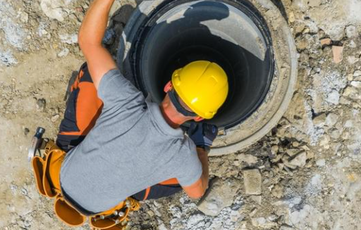 Worker in hard hat and tool belt looking into a manhole on a gravel surface. The setting is outdoors, with construction materials around.