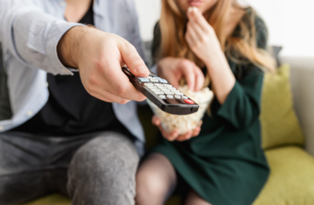Man points a TV remote while a woman eats popcorn on a couch, both watching something with focused attention.