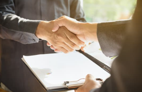 Two people are shaking hands over a clipboard with papers, set in a bright room. The mood is professional and cooperative.