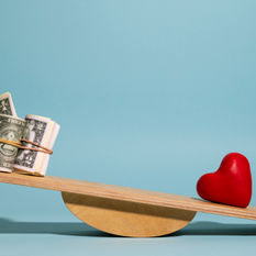 Image of rolled-up dollar bills and a red heart on a wooden balance beam (credit, freepik)