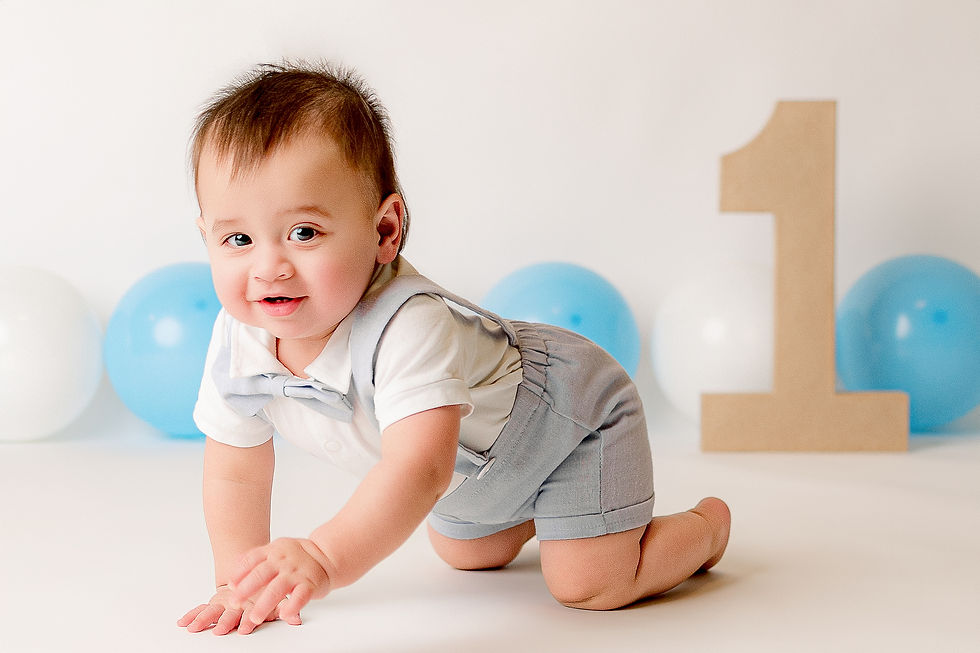 Smiling baby crawls in a cute outfit with a bowtie. Light blue and white balloons, and a large cardboard "1" in the background. Happy mood.