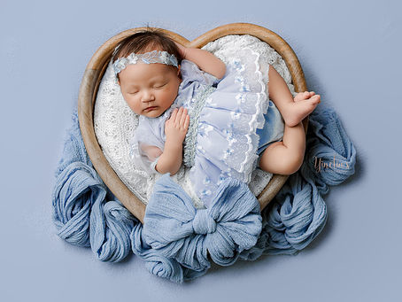 Newborn baby girl Sasha, 23 days old, in fall-themed colors with a matching floral headband, wearing a blue Cinderella dress in a wooden heart bowl, and posed on a newborn table during a professional photography session at Yinelias Photography in Missouri City, TX.