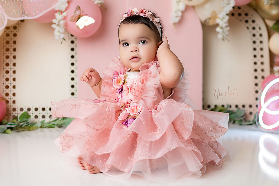 Baby in a pink butterfly dress and floral headband sits on a white floor, touching her ear. Floral and butterfly decor in background.