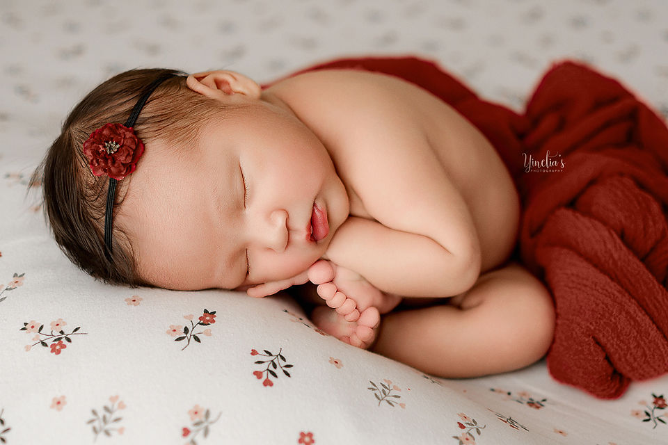 Sleeping baby on floral blanket, wrapped in red fabric. Wears a red flower headband. Peaceful mood. Text: Yinchia's Photography.