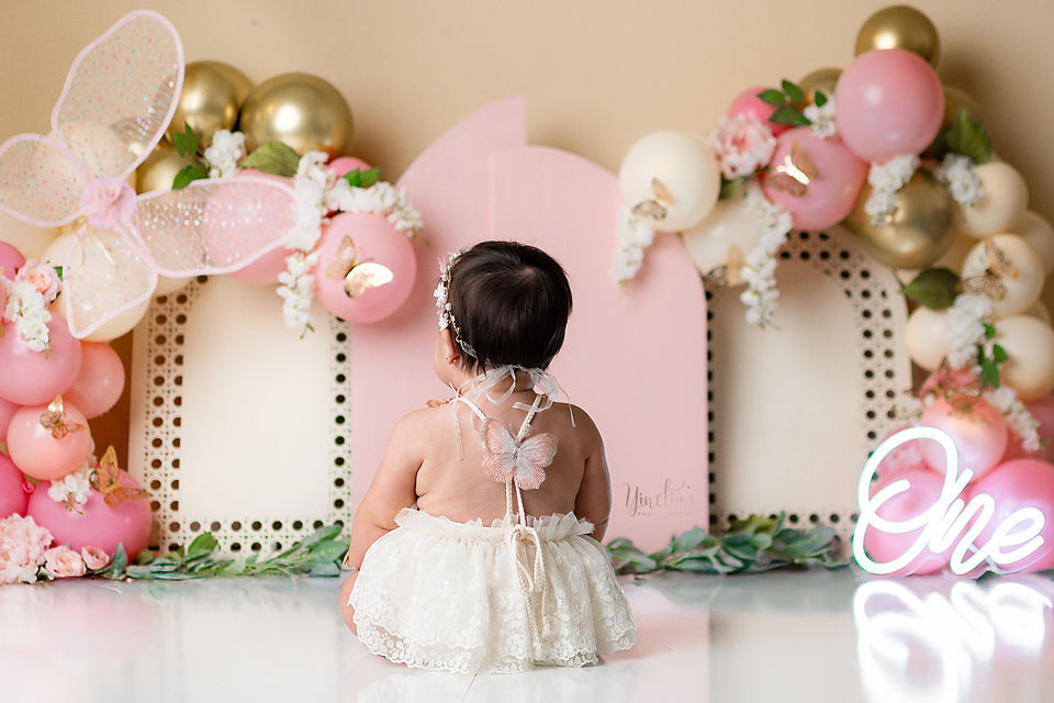 Baby in lace dress with butterfly wings sits among pink, white, and gold balloons. "One" neon sign on right; festive setting.