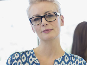 A Caucasian woman with short blonde hair and glasses, smiling at the camera against a neutral background