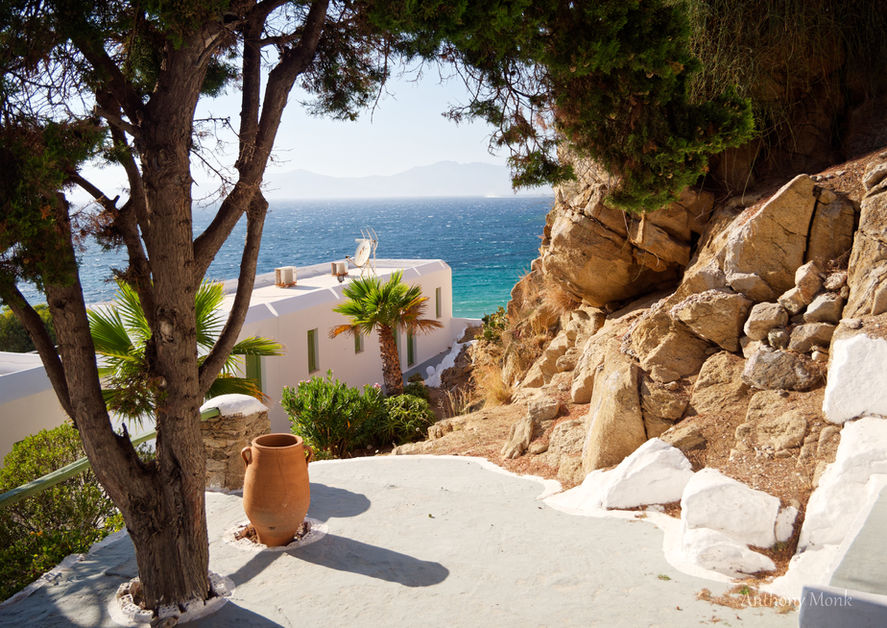 Full midday sun shining on Greek Mediterranean scene. Palm trees and pot in the foreground. Aegean sea in the background and island horizon.