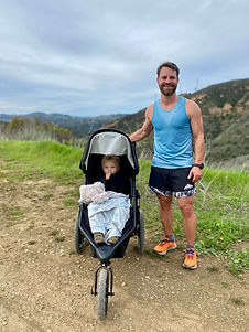 Chris is standing in the Santa Ana mountains with his son in his running stroller. His son has a blanket, stuffed animal and is sucking his thumb.