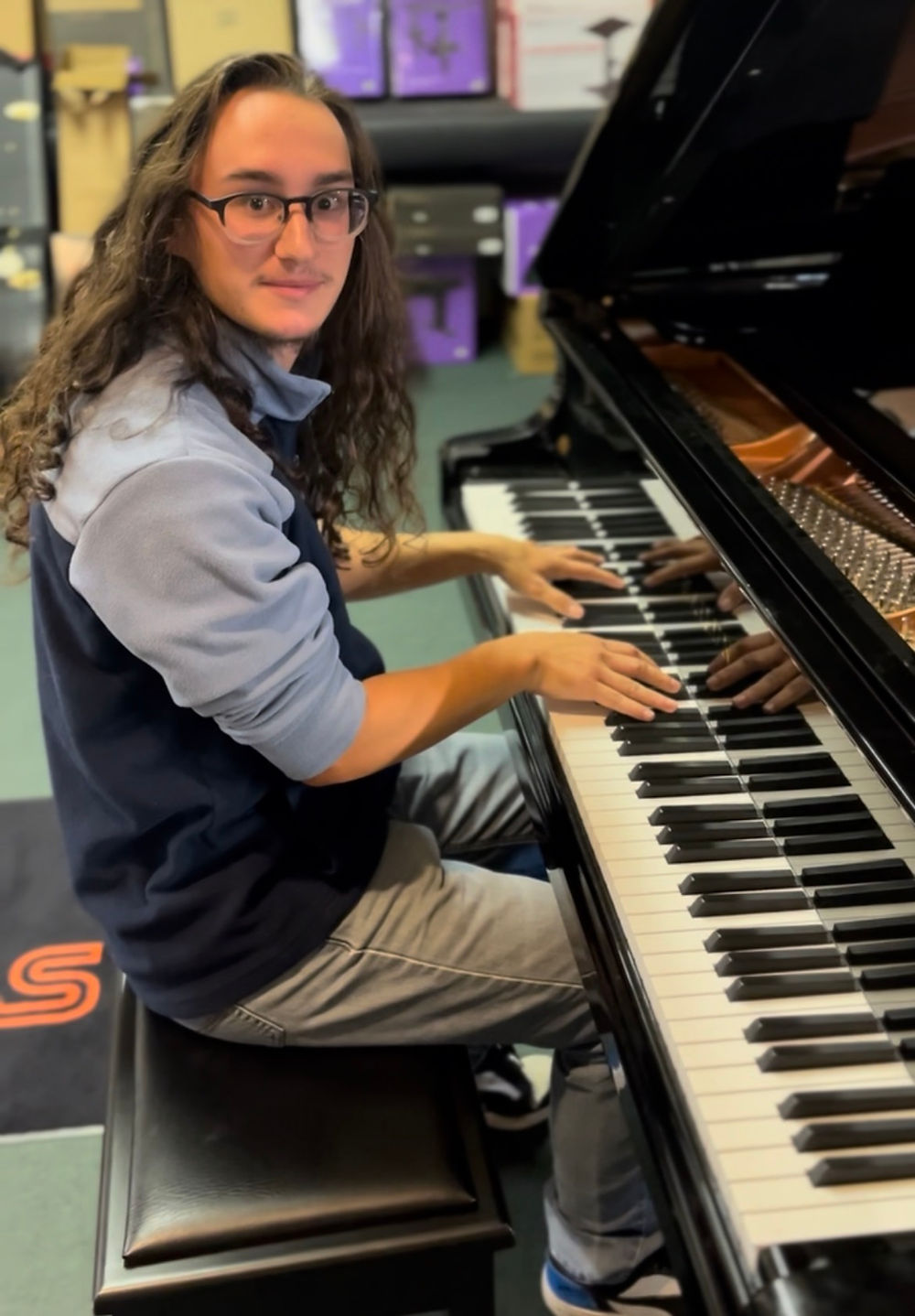 Young man with long hair and glasses plays grand piano, looking forward.