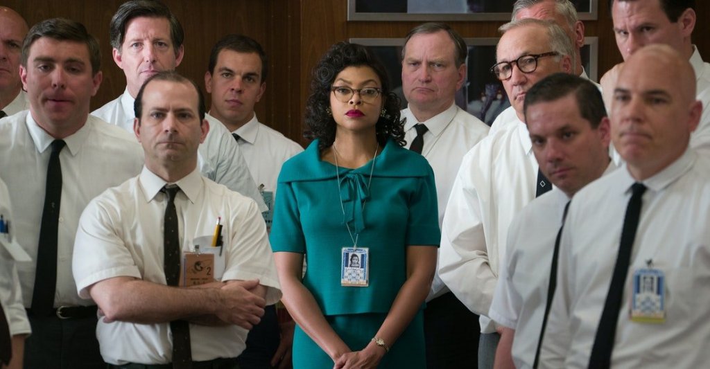 A screen still from Hidden Figures, featuring Katherine Goble Johnson and group of white men in suits, all looking intensely at something.