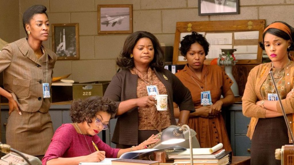 A screen still from Hidden Figures, featuring Katherine Goble Johnson, Mary Jackson, and Dorothy Vaughn standing around a small office with their coworkers. They all look concerned, except for Johnson, who is writing.