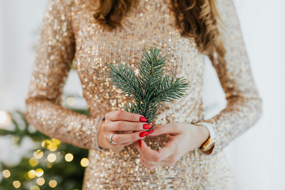 Woman in a sequined gold dress holds a sprig of pine. Red nails, subtle bokeh lights in the background create a festive mood.