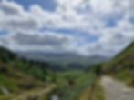 Mountain landscape with a rocky path winding through green hills, trees dotting the slopes, under a cloudy blue sky. Peaceful and scenic.