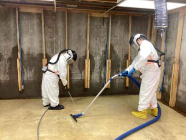 Two people in protective suits clean a concrete basement with vacuum hoses. The mood is diligent. The room has unfinished wood beams.