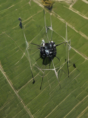 A drone-like machine flies over vast, green agricultural fields with a grid pattern, casting thin shadows. Bright day, no visible text.