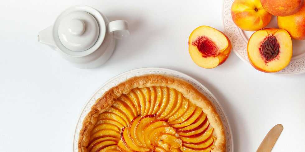 Peach tart with a golden crust on a white plate, surrounded by peaches and a white teapot, set against a light background.